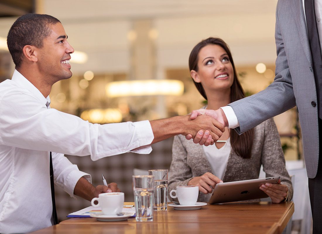 Captives Seminar - Two Professionals Sitting at Table in a Resturant While One is Shaking the Hand of Another Professional Who is Standing by the Table